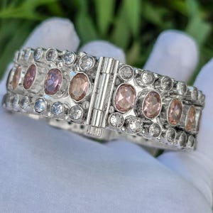 May include: Silver hinged bracelet with oval pink and clear round gemstones. The bracelet has a secure clasp. The bracelet is held against a green background.