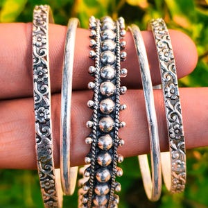 May include: A selection of silver bangles. Two are adorned with floral patterns, and one has a series of small, raised spheres. The bangles are displayed against a blurred green backdrop. The bangles are likely made of sterling silver.
