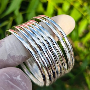 May include: A stack of shiny silver bangles. The bracelets are smooth and reflective, with a polished finish. The bangles are stacked on a light-colored surface, with a blurred green background.