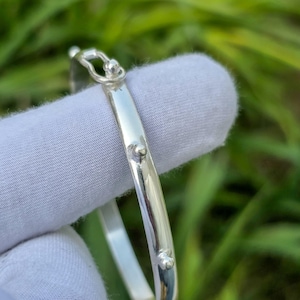 Könnte beinhalten: Ein glänzendes silbernes Armreif-Armband mit Hakenverschluss. Das Armband hat eine glatte, polierte Oberfläche und kleine, dekorative silberne Akzente. Das Armband wird vor einem grünen Hintergrund gezeigt.