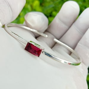 May include: A silver cuff bracelet with a rectangular, deep red gemstone. The bracelet has a simple, elegant design with a polished finish. The gemstone is set in a bezel setting, adding a touch of colour and sophistication to the jewellery.