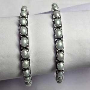 May include: Two silver bangles, each adorned with a series of oval, pale-coloured stones. The bangles are displayed on a white surface. The stones are evenly spaced and resemble pearls.