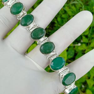 May include: A silver bracelet featuring oval-cut, vibrant green gemstones. The gemstones are set in individual silver bezels and linked together. The bracelet is displayed against a green background.