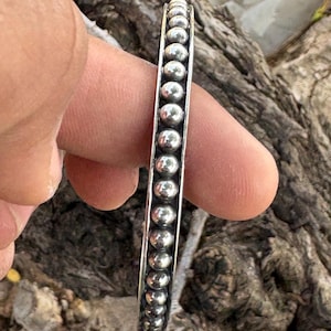 May include: A silver cuff bracelet featuring a single row of polished, spherical beads set between two thin, raised metal borders. The bracelet has a metallic, reflective finish and a uniform, repeating circular pattern along its entire length.