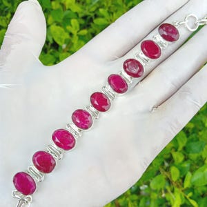 May include: A silver bracelet featuring oval-cut, vibrant red gemstones. The gemstones are set in individual silver bezels and linked together. The bracelet is displayed against a white background, showcasing the jewelry's design and color.
