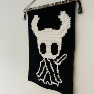 Könnte beinhalten: Schwarz-weißer Wandbehang aus Häkelarbeit mit einem Charakterdesign. Das Design ist eine weiße Figur auf schwarzem Hintergrund. Der Wandbehang wird an einer Holzstange mit schwarzem Faden und Quasten aufgehängt.