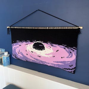 May include: A black and purple crocheted wall hanging depicting a black hole and galaxy. The artwork is suspended from a wooden dowel with black string. The color palette includes black, purple, white, and lavender.