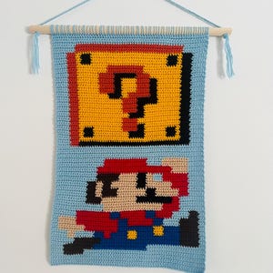 Może przedstawiać: Jasnoniebieski, szydełkowy gobelin ścienny z pikselowym wzorem postaci z gry wideo Mario skaczącego pod żółtym blokiem ze znakiem zapytania. Zawieszony na drewnianym drążku z niebieskimi frędzlami z przędzy.