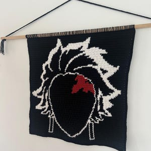 Könnte beinhalten: Ein schwarzer Wandbehang aus Häkelarbeit mit einem weißen und roten Charakterdesign. Das Design ist eine stilisierte Silhouette mit herzförmigem Gesicht und stacheligem Haar. Der Wandbehang ist an einem Holzstab mit einer schwarzen Kordel aufgehängt.