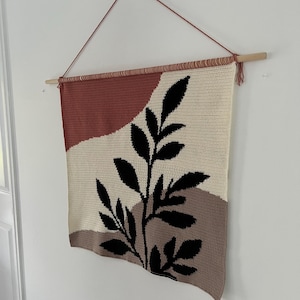Könnte beinhalten: Ein rechteckiger, handgefertigter Wandbehang mit botanischem Design. Das Stück hat einen cremefarbenen Hintergrund mit einem schwarzen Blättermuster und abstrakten Formen in Braun- und Rottönen. Es wird an einer Holzstange mit einer roten Schnur aufgehängt.