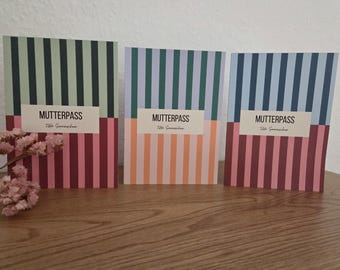 May include: Three rectangular booklets with vertical striped covers in varying color combinations. Each cover features a white rectangular label with the black text MUTTERPASS and Tilda Sonnenschein. The booklets are displayed side by side.