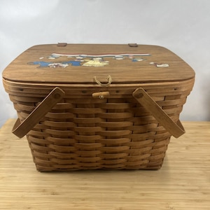 May include: A brown woven picnic basket with a wooden lid. The lid features a painted design of cartoon figures. The basket has two wooden handles and a wooden latch. The basket is rectangular and has a woven pattern.