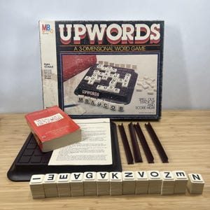 May include: A vintage Upwords board game set. The box is black and white with red accents, and the game board features a grid for stacking letter tiles. Includes a dictionary, letter tiles, and game instructions.