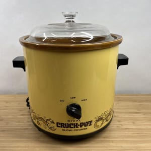 May include: A yellow Rival Crock-Pot slow cooker with a clear glass lid and black side handles. The front features a black control knob with Off, Low, and High settings, along with brown line art illustrations of food and the text Rival Crock-Pot.