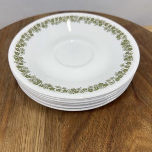 以下が含まれることがあります： 縁に緑色の花柄模様が入った白いソーサーのスタック。ソーサーは木製の表面に積み重ねられています。ソーサーは、ヴィンテージ食器セットの一部である可能性があります。
