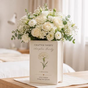 Puede incluir: Un arreglo floral de rosas blancas, paniculata y otras flores blancas en un jarrón crema con forma de libro. El jarrón tiene el texto "CHAPTER THIRTY" y una ilustración de clavel. El arreglo está sobre una mesa de madera.