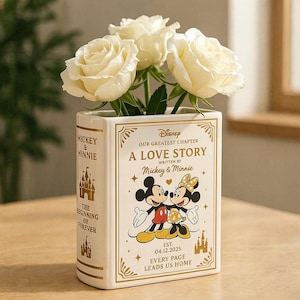 May include: A white ceramic book-shaped vase featuring gold text and illustrations of Mickey and Minnie Mouse. The front reads A LOVE STORY, OUR GREATEST CHAPTER, and EST. 04.12.2025. Three cream-colored roses are arranged inside the rectangular vessel.