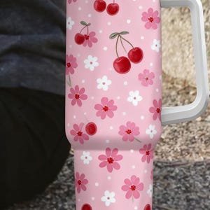 May include: Pink tumbler with a white handle, lid, and straw. The tumbler is decorated with a pattern of red cherries, pink flowers, and white flowers. The top of the tumbler has a gold band.