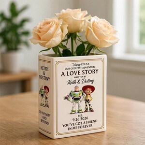 Può includere: Un vaso rettangolare in ceramica bianca a forma di libro, con i personaggi di Toy Story della Disney Pixar, Buzz Lightyear e Jessie. Il testo recita A LOVE STORY, WRITTEN BY KEITH & DESTINY, EST. 9.26.2026, YOU'VE GOT A FRIEND IN ME FOREVER.