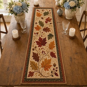 May include: A rectangular quilted table runner featuring a cream-colored center panel with an embroidered pattern of autumn leaves in shades of red, orange, and green, accented by small acorns and swirling vines, bordered by a dark green trim.