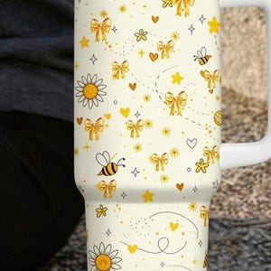 May include: A white travel mug with a handle, decorated with a pattern of yellow bows, bees, hearts, and daisies. The mug has a stainless steel band and a white lid. The design is set against a cream-coloured background.
