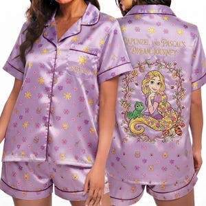 May include: A lavender satin pajama set featuring a short-sleeved button-down shirt and matching shorts. The fabric has a repeating pattern of suns, flowers, and honey pots. The back displays Rapunzel and Pascal with the text Rapunzel and Pascal's Dream Journey.