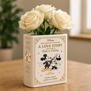 May include: A white ceramic book-shaped vase featuring gold text and illustrations of Mickey and Minnie Mouse. The front reads Disney Our Greatest Chapter A Love Story Written By Keith & Destiny Est. 9.26.2026 Every Page Leads Us Home.