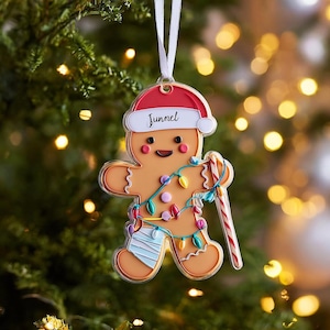 May include: Gingerbread man Christmas ornament with a red Santa hat and the name "Junnel". The ornament has a candy cane, a string of colorful lights, and a bandage on its leg. It hangs from a white ribbon.