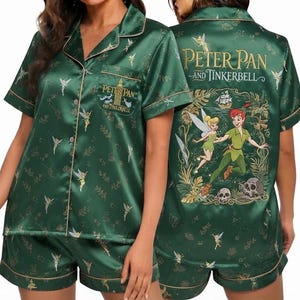 Puede incluir: Un conjunto de pijama de satén verde oscuro que incluye una camisa de manga corta con botones y pantalones cortos a juego. La tela tiene un patrón repetido de Campanilla. El bolsillo de la camisa muestra el texto "Peter Pan and Tinkerbell" con el Big Ben, y la parte posterior presenta un gráfico grande de Peter Pan y Campanilla con el texto "Peter Pan and Tinkerbell".