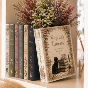 Op de afbeelding: Een verzameling decoratieve boeken met bloemmotieven en titels als "Pride and Prejudice" en "Rebecca". Een groot boek met de titel "Sophia's Library" toont een zwarte kattenillustratie en de tekst "Stories live forever in the heart."