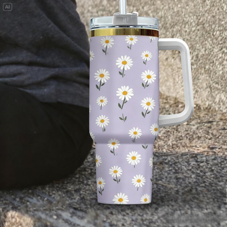 May include: A large, insulated tumbler with a white handle and a clear lid. The tumbler is light purple and covered in a pattern of white daisies with yellow centers and green stems. The rim of the tumbler has a gold band.