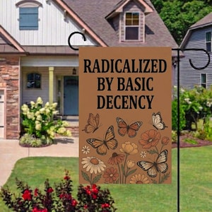 May include: A brown garden flag displaying the text "RADICALIZED BY BASIC DECENCY" in black. The flag features illustrations of butterflies and flowers in brown, orange, and white. The flag is hanging in a front garden.