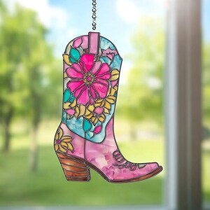 Puede incluir: Un atrapasoles de bota de vaquero de vidriera rosa con un diseño floral. La bota presenta una gran flor rosa, detalles amarillos y turquesas y un tacón marrón. Cuelga de una cadena plateada.