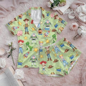 Puede incluir: Un conjunto de pijama verde claro que consta de una camisa de manga corta con botones y pantalones cortos a juego. La tela presenta un estampado integral de personajes de Toy Story, incluidos Woody, Buzz Lightyear, Jessie, Rex, Hamm y los aliens.