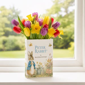 Könnte beinhalten: Eine rechteckige Vase mit dem Peter Rabbit-Design von Beatrix Potter, gefüllt mit bunten Tulpen und Narzissen. Die Vase zeigt den Text "Peter Rabbit by Beatrix Potter". Die Blumen sind rot, gelb und lila.