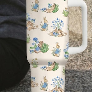May include: A white travel mug featuring a repeating pattern of rabbits in blue jackets, flowers, and greenery. The mug has a white handle, lid, and straw. The mug is approximately 10 inches tall.