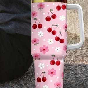 May include: A pink tumbler with a white handle, lid, and straw. The tumbler is decorated with a pattern of red cherries, pink flowers, and white polka dots. The rim has a gold accent.