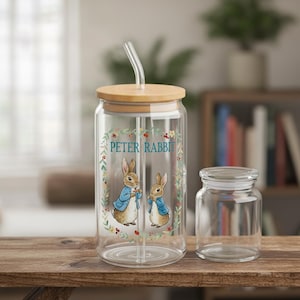 May include: A clear glass tumbler with a wooden lid and a curved straw. The tumbler features a Peter Rabbit design with two rabbits wearing blue jackets. The words "PETER RABBIT" are printed on the glass, surrounded by a floral wreath. A small, clear glass jar sits next to the tumbler.