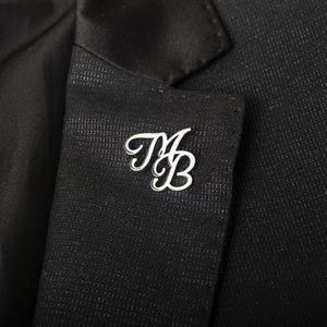 May include: A silver lapel pin, featuring the intertwined cursive initials "MB", is attached to the black fabric of a suit jacket. This decorative pin is a stylish accessory, adding a touch of elegance to formal attire.