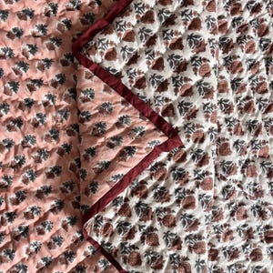 May include: A reversible quilt with a floral pattern. One side features a peach background with dark gray floral designs, while the other side has a white background with brown floral designs. The quilt is trimmed with a burgundy border.