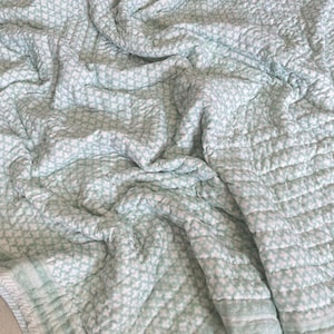 Puede incluir: Una manta acolchada suave con un patrón floral repetitivo en blanco y verde claro. La manta tiene una apariencia texturizada y está doblada, mostrando los detalles de las costuras. La paleta de colores general es una estética pastel relajante.