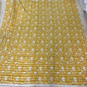 Könnte beinhalten: Eine gelbe Steppdecke mit weißem Blumenmuster. Die Steppdecke ist gefaltet und zeigt die gesteppte Textur und den weißen Rand. Das florale Design ist gleichmäßig über dem gelben Stoff verteilt und erzeugt ein optisch ansprechendes Textil.