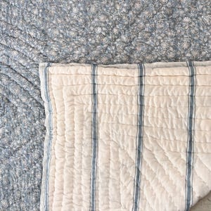 Könnte beinhalten: Ein gestepptes Textil mit zwei verschiedenen Mustern. Ein Bereich zeigt einen dichten, komplexen Blumendruck in gedämpftem Blau und Weiß. Der andere Bereich ist ein cremefarbener Stoff mit vertikalen blauen Streifen und einer gesteppten Gitterstruktur.