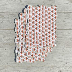 Peut inclure: Quatre dessous de verre en tissu blanc avec un motif floral orange et marron répété. Les dessous de verre ont un bord festonné avec une bordure verte. Ils sont posés sur une surface en bois clair.