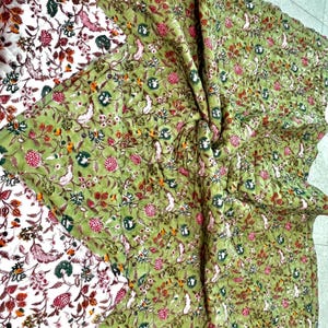 May include: A folded green fabric with a floral pattern featuring pink, white, and brown flowers, along with white birds and green leaves. The fabric is layered over a white fabric with a similar floral design.