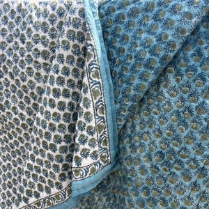 May include: A reversible quilted textile featuring a block-print pattern of small green floral motifs. One side has a white background, while the other side has a light blue background. A border with a repeating botanical design edges the fabric.