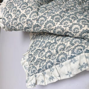 May include: A folded quilt with a floral pattern in shades of blue and white. The quilt features a repeating design of stylized flowers and a ruffled edge. The fabric appears to be soft and comfortable.