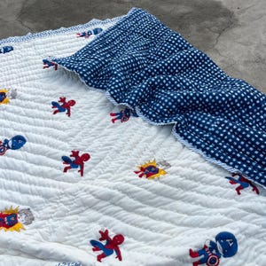 May include: A white quilted blanket featuring a repeating pattern of embroidered superhero characters including Spider-Man and Captain America. The blanket is folded over to reveal a dark blue and white checkered fabric on the reverse side.