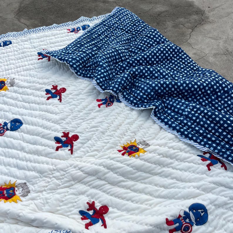 May include: A white quilted blanket featuring a repeating pattern of embroidered superhero characters including Spider-Man and Captain America. The blanket is folded over to reveal a dark blue and white checkered fabric on the reverse side.
