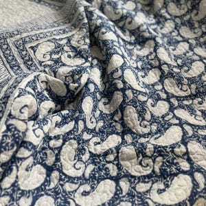 Könnte beinhalten: Eine gefaltete, gesteppte Decke mit einem marineblauen Paisley-Muster auf weißem Grund. Die Decke hat einen dekorativen Rand mit einem floralen Design. Der Stoff ist weich und scheint aus Baumwolle zu sein.
